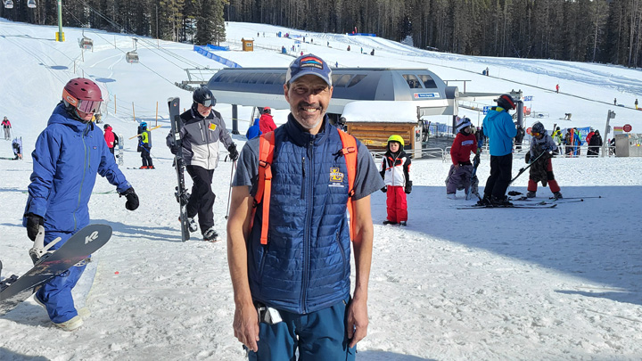 A smiling man in ski attire stands near a ski lift on a snowy mountain.