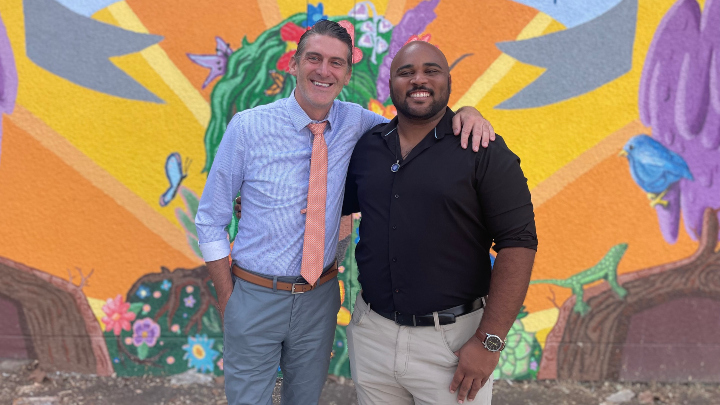 Two smiling men in business attire pose in front of a brightly colored mural.