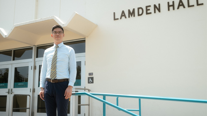 A man in a white shirt and yellow tie stands in front of Lamden Hall.