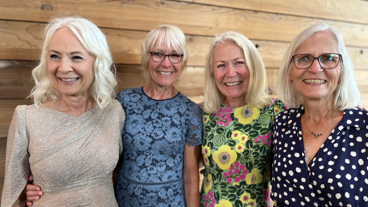 Four women smile standing in front of a wood paneled wall.