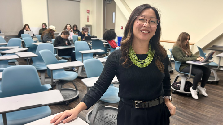 A woman in black smiles while standing at the front of a classroom.