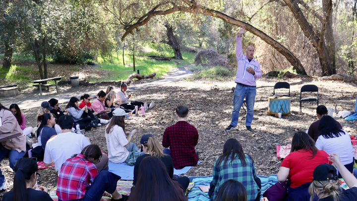 A man in a pink jacket points skyward as he speaks to a group of seated students in the woods.