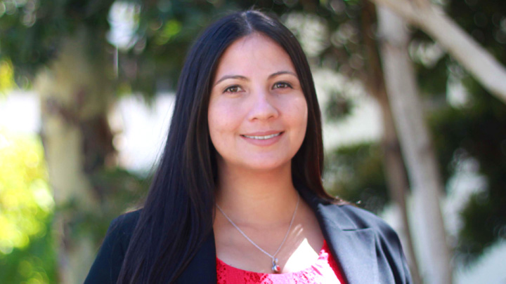 A woman in a black blazer and red shirt smiles in front of a green backdrop.